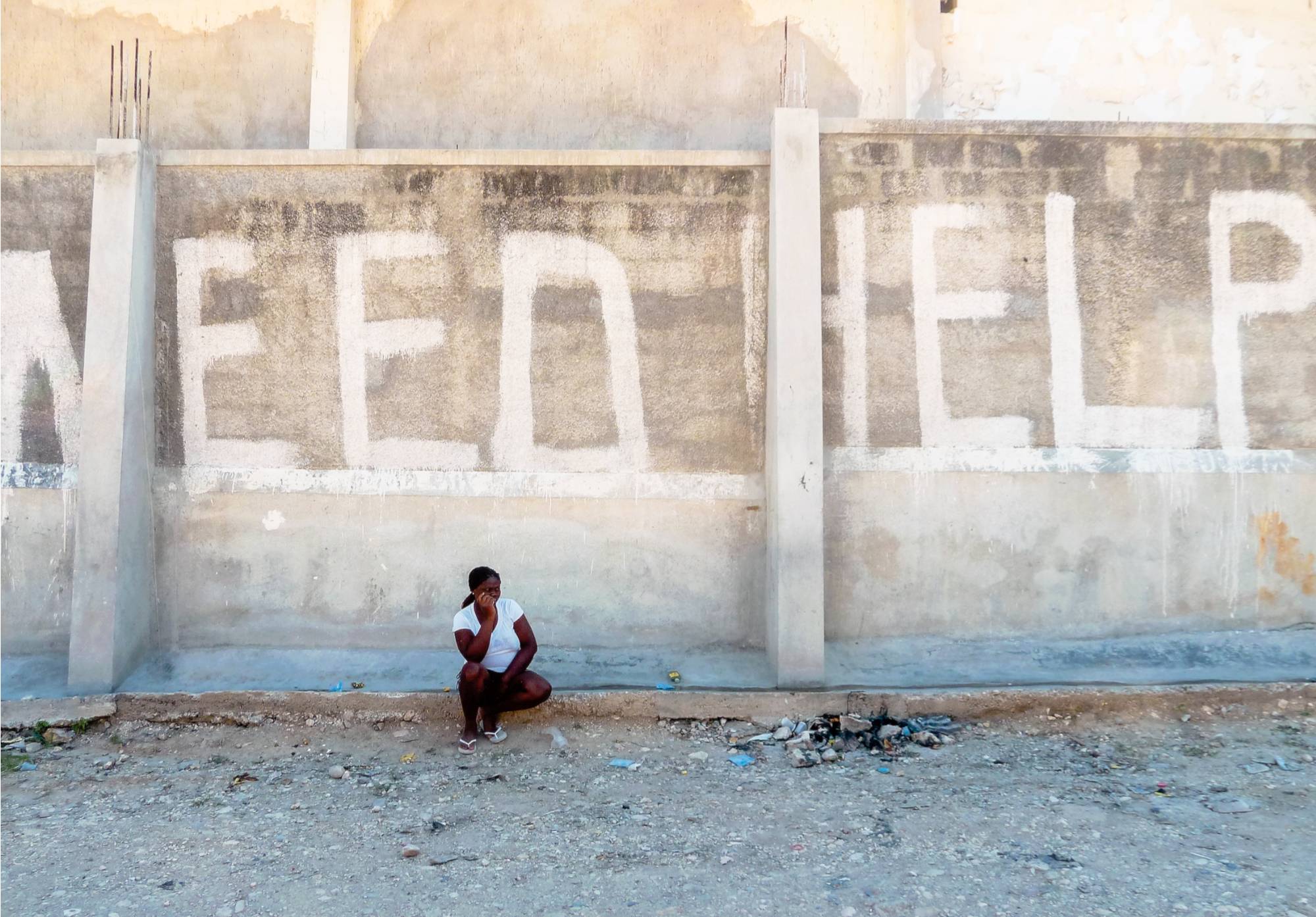 Photograph of a person kneeling near a wall that says "Need Help" in Haiti after an earthquake.